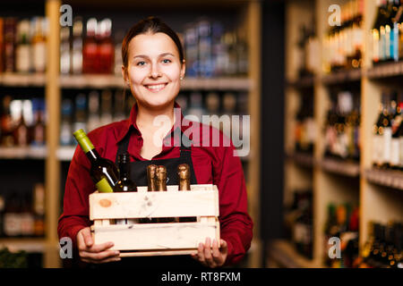 Immagine della donna felice con scatola di legno con bottiglie nelle sue mani nel negozio del vino Foto Stock