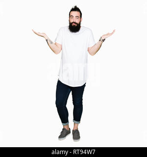 Tanga giovane uomo con i capelli lunghi e la barba che indossa casual t-shirt bianco clueless e espressione confusa con le braccia e le mani alzate. Concetto di dubbio. Foto Stock