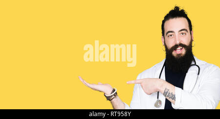 Medico con capelli lunghi che indossa medical e il cappotto di uno stetoscopio stupiti e sorridente alla fotocamera durante la presentazione con la mano e puntando con il dito. Foto Stock