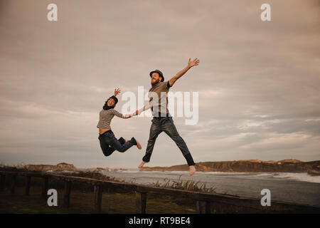 Giovane donna felice e uomo in cappelli tenendo le mani e divertirsi sul sedile sulla costa vicino mare ondeggiante e cielo nuvoloso Foto Stock