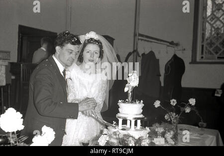 Degli anni Cinquanta, gli sposi si tengono per mano il taglio della torta di nozze insieme al loro ricevimento in una sala della chiesa, Inghilterra, Regno Unito. Foto Stock