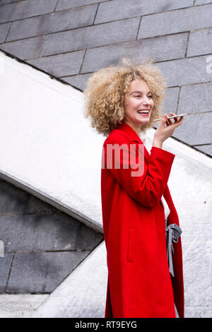 Ritratto di ridere donna bionda con boccoli indossando cappotto rosso parlando al telefono cellulare Foto Stock