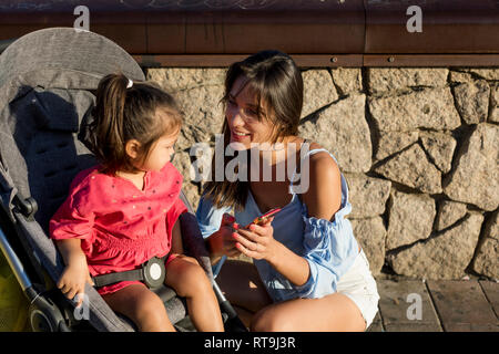 Madre inginocchiato accanto alla figlia, seduti nella PRAM, guardando a smartphone Foto Stock