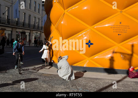 Un ciclisti gira l'angolo e guarda a un modello di moda essere fotografati contro il temporaneo di accaparramento di rinnovamento del marchio di lusso di Louis Vuitton a New Bond Street, il 25 febbraio 2019, a Londra, in Inghilterra. Foto Stock