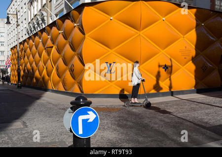Una signora su un scooter elettrico passa il temporaneo di accaparramento di rinnovamento del marchio di lusso di Louis Vuitton a New Bond Street, il 25 febbraio 2019, a Londra, in Inghilterra. Foto Stock
