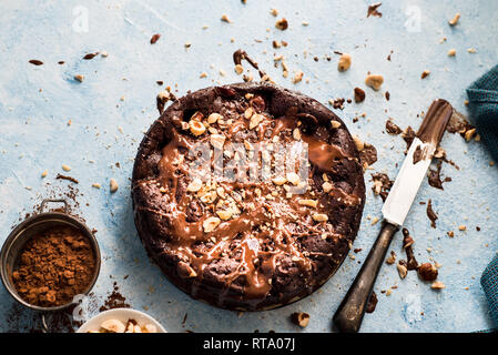 Una deliziosa torta al cioccolato con glassa di cioccolato e nocciole su un blu sullo sfondo a trama con un coltello e cacao in una succhieruola Foto Stock