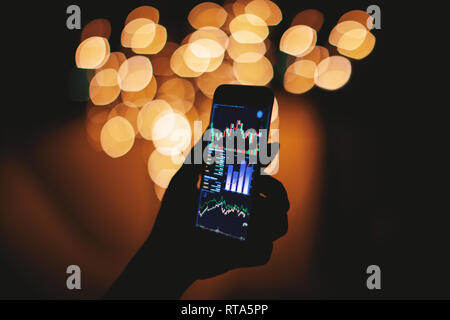 Silhouette di donna mano azienda smart phone al buio con stock trading sul display con luce bokeh sfondo sfocato mobile business concept Foto Stock