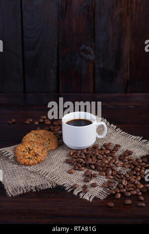 White tazza di caffè con caffè in chicchi vicino e biscotti sul tessuto di lino sul vintage scuro dello sfondo. Foto Stock