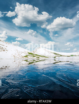 Congelati lago di montagna con il blu ghiaccio e crepe sulla superficie. Il pittoresco paesaggio invernale con colline innevate sotto un azzurro cielo nuvoloso Foto Stock