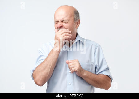 Bald ritirato l'uomo sente assonnato, sbadigli come si sente stanco, apre la bocca ampiamente, vestito in maglietta blu. Stanchezza e concetto di pensione Foto Stock