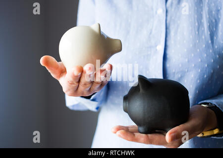 Persona detiene due piggy banche, il bianco è più piccolo e leggero, nero è più grande e più pesante, come il risparmio di debito vs Foto Stock