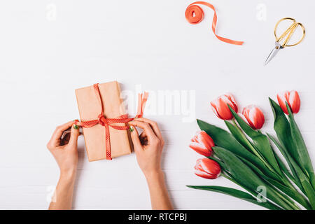 Vista superiore della femmina mani prua di legatura sulla confezione regalo accanto al bouquet di tulipani rossi e forbici d'oro bianco su un tavolo di legno. Foto Stock