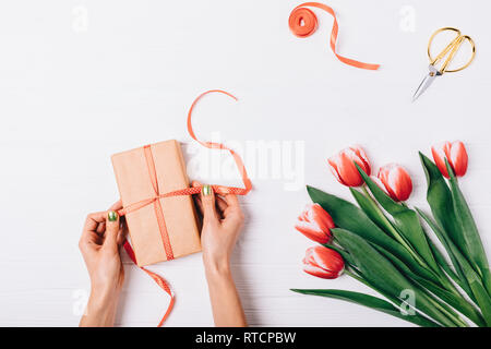 Vista dal di sopra della donna con le mani in mano la legatura della confezione regalo con nastro prossima a bouquet di tulipani rossi e forbici d'oro bianco su un tavolo di legno. Foto Stock