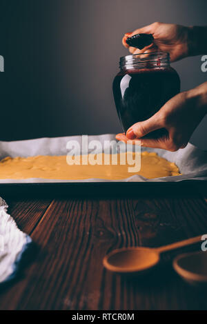 Giovane donna mettendo inceppata sul foglio laminato di impasto per cuocere biscotti fatti in casa. Close-up di donna mani tenendo il vaso e un cucchiaio di confettura di ribes. Foto Stock