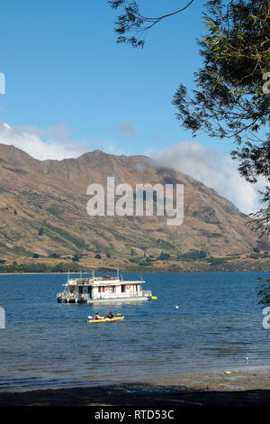 Persone in giallo kayak sul lago Wanaka da una casa galleggiante su una soleggiata giornata estiva, con montagne Wanaka, Nuova Zelanda Isola del Sud Foto Stock
