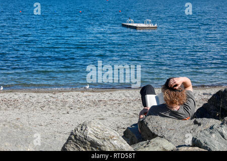 L'uomo la lettura di un libro sotto il sole seduti sulle rocce a Wanaka spiaggia sulla riva del Lago Wanaka Nuova Zelanda Isola del Sud Foto Stock