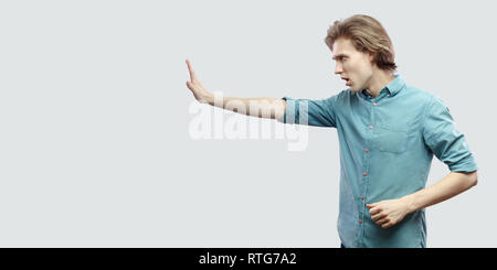 Profilo vista laterale ritratto di grave bello capelli lunghi biondo giovane uomo in blu T-shirt casual in piedi con la mano di arresto gesto segno. piscina studio s Foto Stock