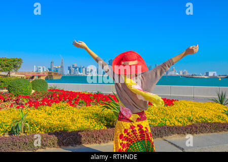Carefree donna con ampia hat gode di aiuole fiorite e alberi in fiore sulla Corniche promenade della baia di Doha in Qatar. Stile di vita turistica caucasico guardando Foto Stock
