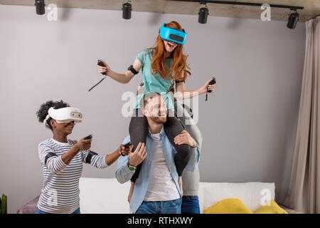 Felice i capelli rossi donna in realtà virtuale auricolare è a cavallo trasportato su il suo fidanzato, circondato da razza mista agli amici a casa. Dating, romantico rel Foto Stock