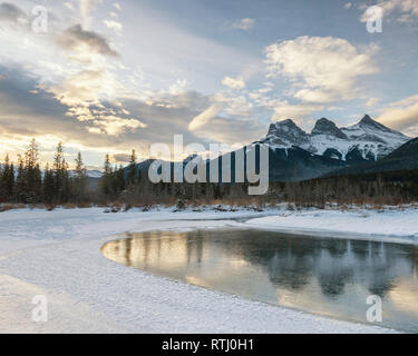 Coperta di neve Tre Sorelle Picchi da poliziotto Creek dopo l'alba si riflette in una parzialmente congelato il Fiume Bow, vicino a Canmore, Alberta, Canada Foto Stock