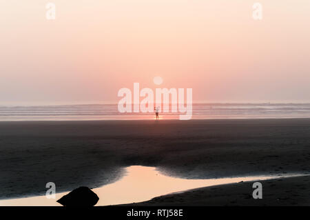 Un surfista solitario che esce con il mare, stagliano contro il sole di setting, Saunton, Devon, Regno Unito. Foto Stock