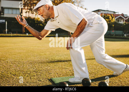 Uomo anziano giocando una partita a bocce. Senior uomo in hat getta a bocce in piedi in posizione in un prato. Foto Stock