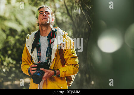 Ritratto di un uomo in giacca e zainetto a camminare in una foresta in possesso di una fotocamera reflex digitale. L'uomo ad esplorare una foresta catturare la bellezza in un digital arrivava Foto Stock