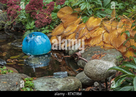 Rocce levigate e denso fogliame, compresi arancio brillante hosta, circondare un piccolo laghetto in giardino che ha un giro, ceramica blu acqua gorgogliatore (autunno). Foto Stock