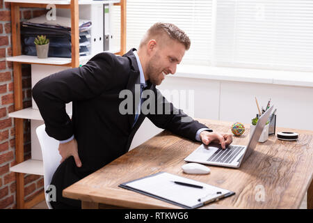 Bel Giovane Maschio avente Executive Backpain lavorando sul computer portatile in ufficio Foto Stock