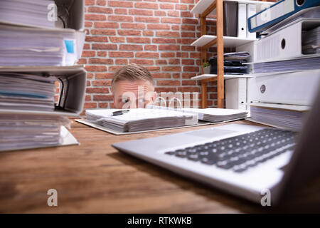 Close-up di imprenditore di nascondersi dietro la scrivania con pila di cartelle e laptop Foto Stock