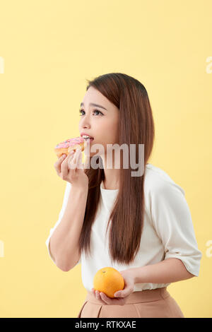 Concetto di dieta. Bella giovane donna asiatica holding arancione e ciambella su sfondo giallo. La scelta tra il cibo spazzatura con alimenti sani. Foto Stock