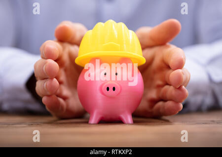 Close-up di mano di una persona di colore rosa di protezione Salvadanaio con Casco giallo Foto Stock