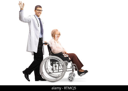 Lunghezza piena ripresa di profilo di un giovane sorridente maschio positivo medico sventolando e spingendo un senior disabili donna in una sedia a rotelle isolato su bianco backgrou Foto Stock