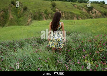 Estate a piedi su un burrone verde, una giovane slim ragazza carina con capelli lunghi marrone in un abito giallo sundress, gode la vita sul campo con fiori d'estate Foto Stock