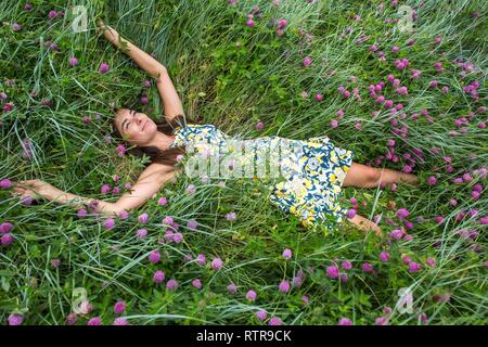 Estate a piedi su un burrone verde, una giovane slim ragazza carina con capelli lunghi marrone in un abito giallo sundress, gode la vita sul campo con fiori d'estate Foto Stock