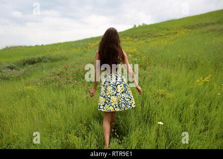 Estate a piedi su un burrone verde, una giovane slim ragazza carina con capelli lunghi marrone in un abito giallo sundress, gode la vita sul campo con fiori d'estate Foto Stock