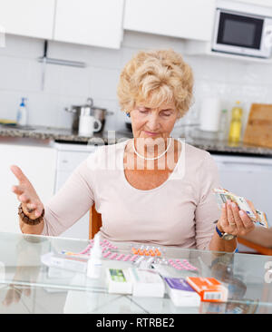 Senior donna contando la sua spesa per pillole seduta in cucina Foto Stock