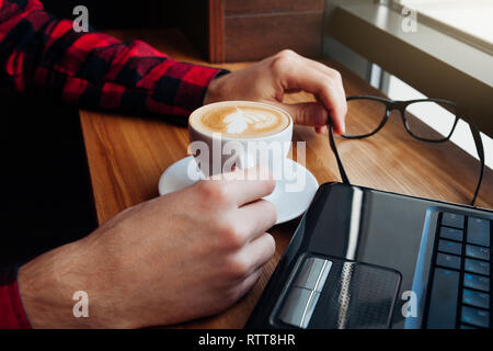 Giovane imprenditore gode di caffè nella caffetteria usando un computer portatile nella parte anteriore della finestra Foto Stock