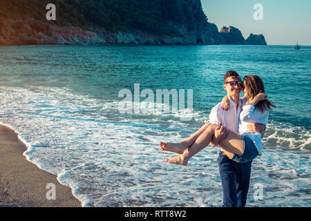 Giovane uomo felice tiene la sua ragazza a braccia sulla spiaggia tropicale al mattino Foto Stock