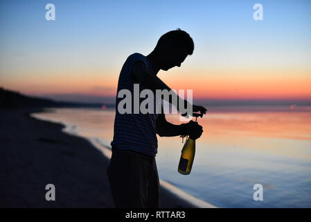 Una silhouette di un ragazzo di apertura di una bottiglia di un vino bianco con un cavatappi. Una notte d'estate sulla spiaggia del Mar Baltico. Foto Stock