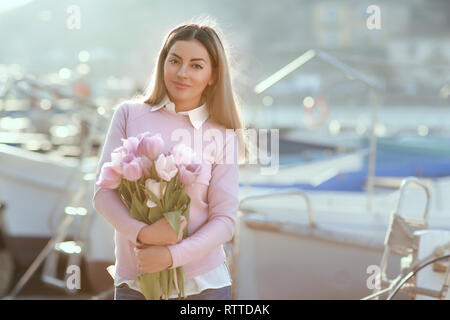 Bella ragazza in primavera o in autunno, in riva al mare nella baia di barca. Con un bouquet di fiori nelle sue mani e serrati eleganti jeans e un maglione rosa Foto Stock