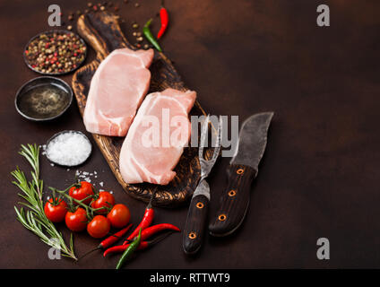 Materie lombo di maiale braciole sul vecchio vintage tagliere con coltello e forchetta sulla scheda arrugginito. Sale e pepe con rosmarino fresco e olio con pepe e toma Foto Stock