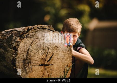 Funny kid caucasica nel capo di nascondersi dietro un log con pistola giocattolo in mani. Ragazzo giocando con pistola giocattolo all'esterno. Foto Stock