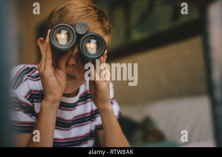 Piccolo Ragazzo che guarda fuori dalla finestra con il binocolo. Ragazzo che guarda attraverso il binocolo dal soggiorno. Foto Stock