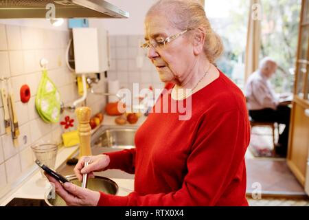 Senior Citizen cuochi una ricetta da Internet, mantiene il telefono cellulare in mano, Renania settentrionale-Vestfalia, Germania Foto Stock