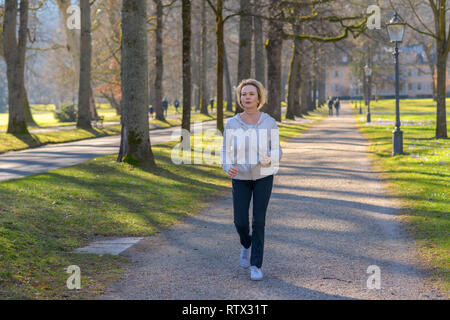 Attraente donna matura prendendo la sua quotidiana jog attraverso un parco boscoso avvicinando la fotocamera lungo un viale alberato in un fitness e salute concetto Foto Stock