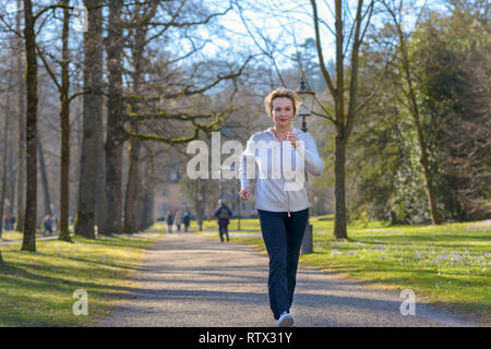 Attraente donna matura prendendo la sua quotidiana jog attraverso un parco boscoso avvicinando la fotocamera lungo un viale alberato in un fitness e salute concetto Foto Stock