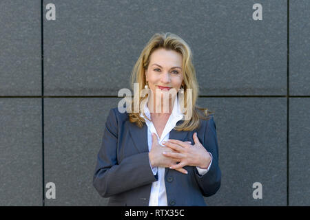 Imprenditrice che mostra la sua sincera riconoscenza e gratitudine stringendo le mani al suo cuore con un sorriso soddisfatto Foto Stock
