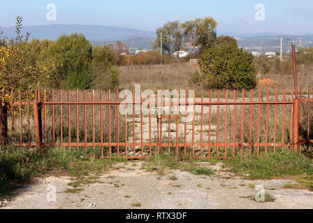 Lungo recinzioni di metallo porte bloccate con catena e lucchetto impedisce l'ingresso di grandi industriali abbandonati complesso circondato con alta erba non tagliata Foto Stock