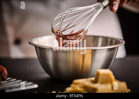 Lo chef prepara il cioccolato fuso in una terrina usando un vecchio frusta in una vista ravvicinata sulla sua mano e utensili Foto Stock